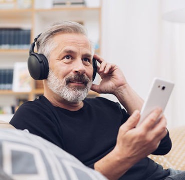 Ein Mann sitzt auf der Couch, hat einen Kopfhörer auf den Ohren und ein Smartphone in der Hand