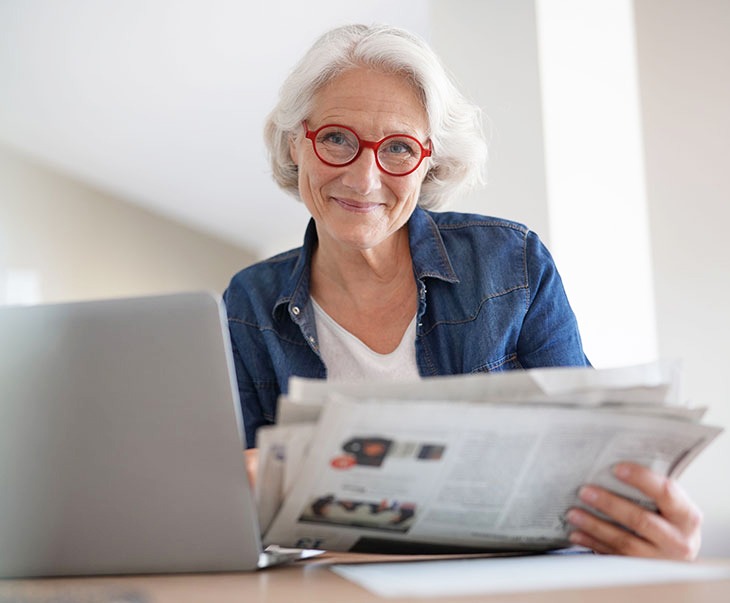 Eine Frau sitzt am Schreibtisch, liest Zeitung und hat einen Laptop vor sich aufgeklappt.